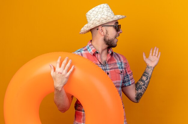 surprised young traveler man with straw beach hat in sun glasses holding swim ring looking at side isolated on orange wall with copy space