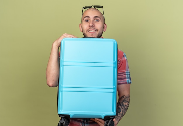 surprised young traveler man with sun glasses holding suitcase isolated on olive green wall with copy space