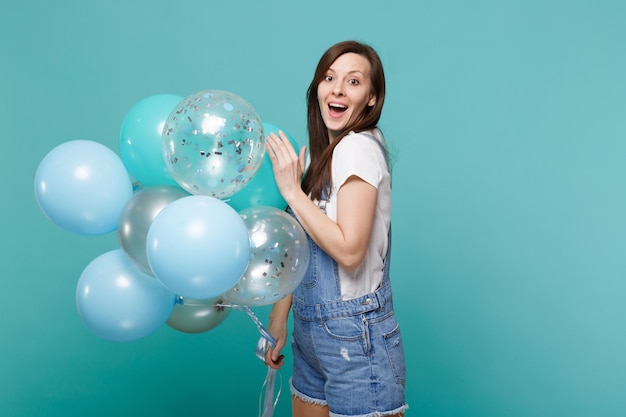 Surprised young woman in denim clothes keeping mouth wide open, celebrating, holding colorful air balloons isolated on blue turquoise wall background. birthday holiday party, people emotions concept.