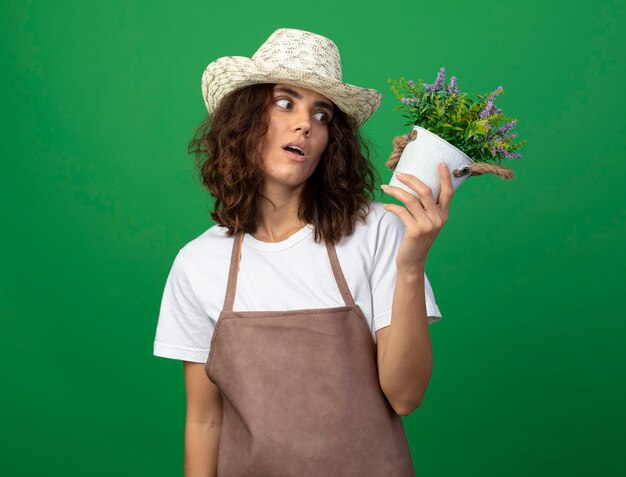 Удивленная молодая женщина-садовник в униформе в садовой шляпе держит и смотрит на цветок в цветочном горшке, изолированном на зеленом
