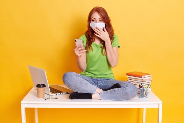 Surprised young woman got news via text message while learning, holding mobile phone and covering her mouth with palm, wear medical mask and casual clothing, sitting on table with crossed legs.