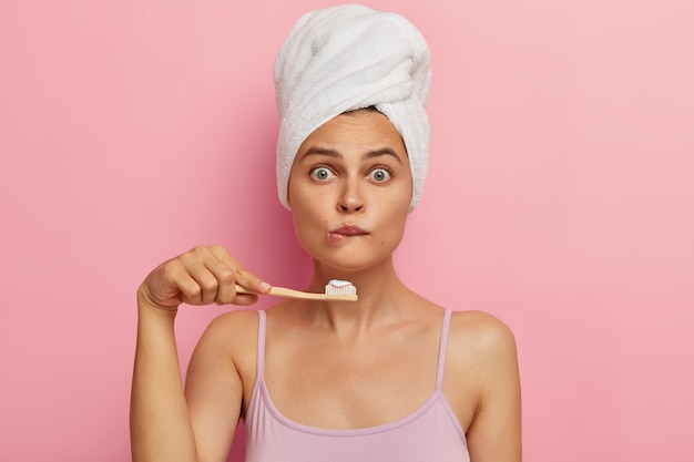 Surprised young woman has morning routine, shocked being short of time, bites lips, holds wooden toothbrush, brushes teeth, wears white towel on head, casual sleeveless shirt