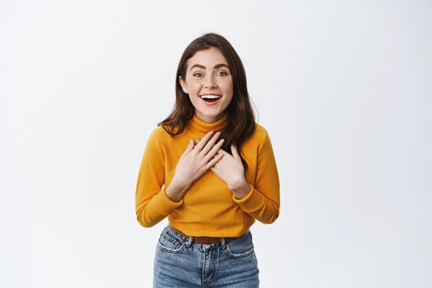 Surprised young woman holding hands on chest amazed and staring at camera recieve surprise gift looking flattered and thankul white background