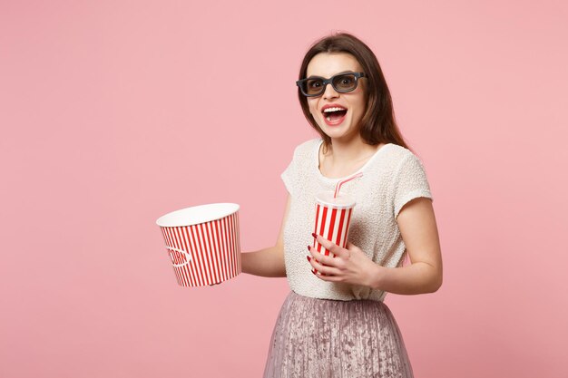 3d 아이맥스 안경을 쓴 놀란 젊은 여성이 파스텔 핑크색 배경에 고립되어 포즈를 취하고 있습니다. 영화 라이프 스타일 개념에서 사람들은 진실한 감정. 복사 공간을 비웃습니다. 콜라 또는 소다의 팝콘 컵 양동이를 잡으십시오.