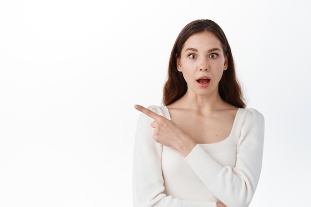 Free photo surprised young woman look in awe pointing index finger aside showing awesome advertisement demonstrate banner wright your text here standing against white background