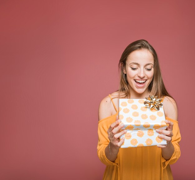 Surprised young woman looking at floral gift box on colored background