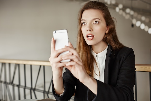 Удивленная молодая женщина смотрит на мобильный телефон, сидя в кафе
