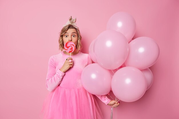 Удивленная молодая женщина выглядит удивленной, когда камера закрывает рот карамельной конфетой в форме сердца, носит праздничное платье, держит кучу гелиевых шаров, изолированных на розовом фоне.