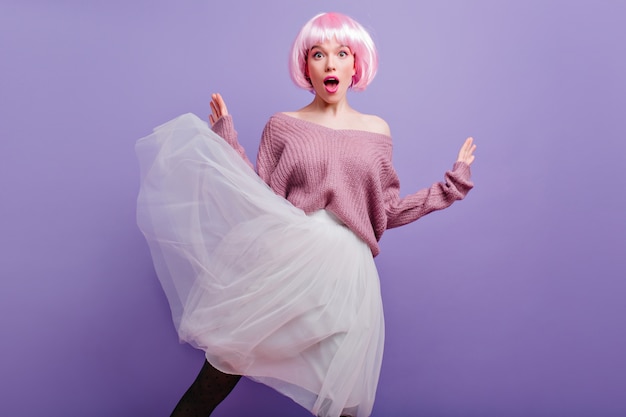 Surprised young woman in lush white skirt posing on purple wall.  fashionable female model in pink peruke expressing amazement.