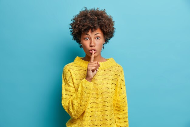 Surprised young woman makes shush gesture asks to keep information in secret shows hush sign has shocked face expression wears casual yellow sweater isolated over blue wall