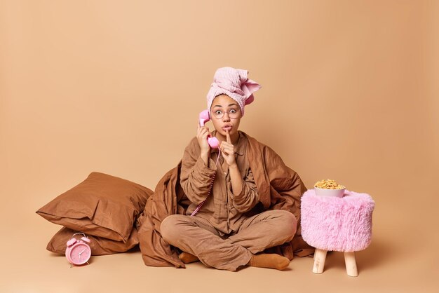 Surprised young woman makes silence gesture talks via stationary telephone wears pajama towel on head covered with blanket shares secret poses crossed legs against brown background has conversation