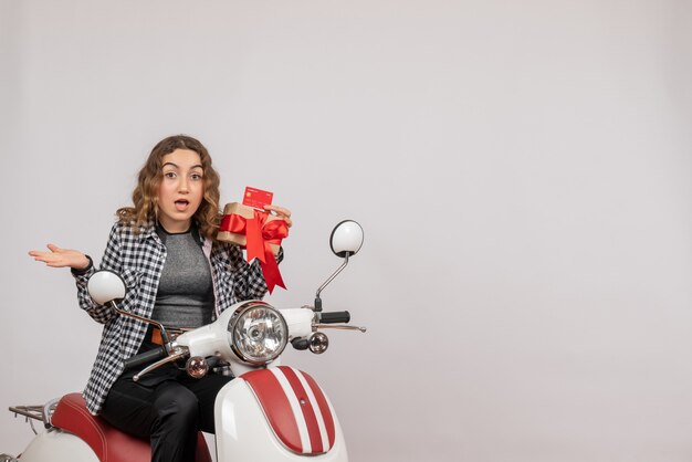 Free photo surprised young woman on moped holding gift and card on grey