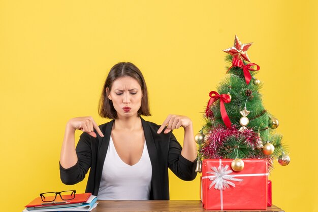 Удивленная молодая женщина, указывающая вниз в костюме возле украшенной рождественской елки в офисе на желтом