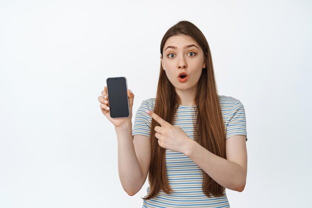 Surprised young woman pointing finger at mobile phone screen, showing smartphone empty and looking amazed at camera on white.