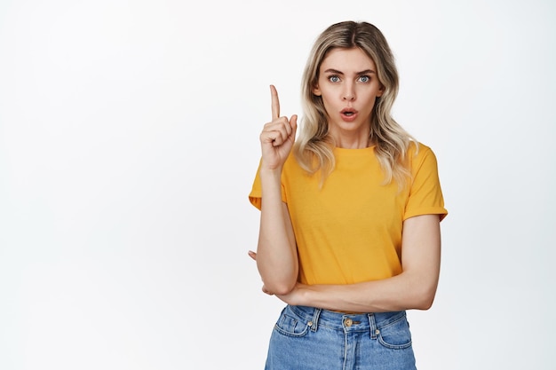 Surprised young woman pointing finger up looking impressed at camera wearing yellow tshirt and jeans white background