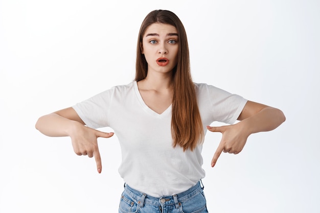 Surprised young woman pointing fingers down, open mouth amazed, raise eyebrows in disbelief, showing something new, banner or logo, standing over white wall