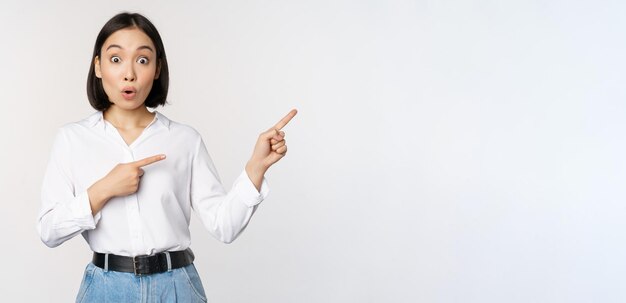 Surprised young woman pointing fingers right Asian girl showing banner and looking enthusiastic interested in advertising standing over white background