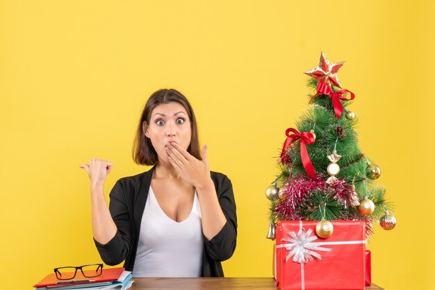 Surprised young woman pointing behind in suit near decorated Christmas tree at office on yellow 