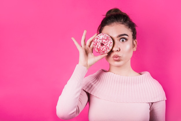 Удивленная молодая женщина надуть губы, закрывая глаза пончиком на розовом фоне