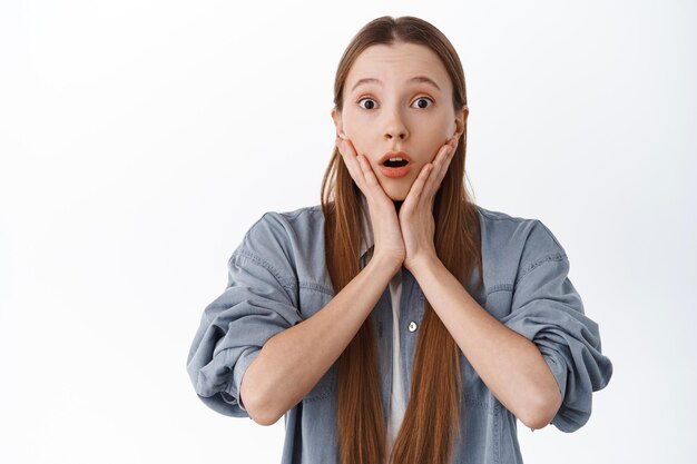 Surprised young woman react to big news, gasping and look in awe, raise eyebrows and open mouth, holding hands on face with shocked expression, white background.