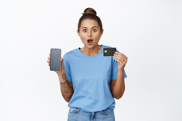 Surprised young woman shows credit card and mobile phone screen. Girl with amazed face shows smartphone interface, shopping app, standing against white background