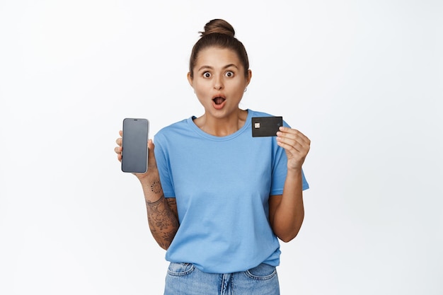 Surprised young woman shows credit card and mobile phone screen. Girl with amazed face shows smartphone interface, shopping app, standing against white background