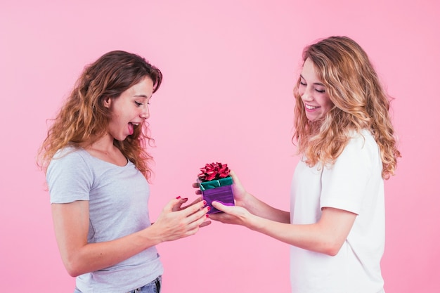 Бесплатное фото Удивленная молодая женщина, принимая подарок от ее друга на фоне розовый