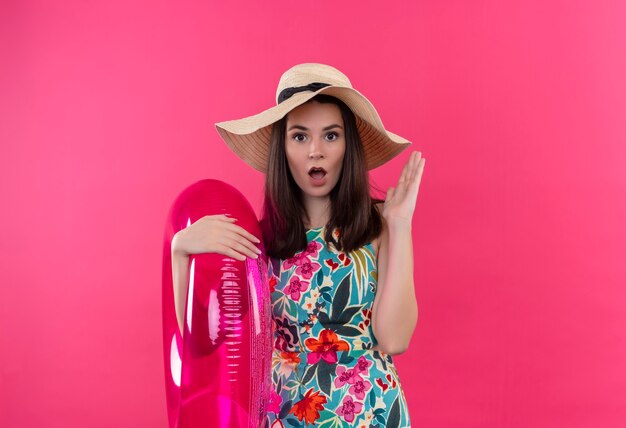 Surprised young woman wearing hat holding swim ring and lifting her hand on isolated pink wall