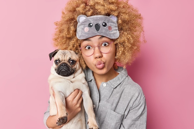 Surprised young woman with curly bushy hair keeps lips folded carries pedigree pug dog spends free time at home with pet wears pajama and sleepmask isolated over pink background Animals concept