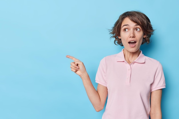 Бесплатное фото Удивленная молодая женщина с короткой прической указывает указательным пальцем с левой стороны на место для рекламы или рекламного текста, привлекая ваше внимание к месту для копирования на синем фоне