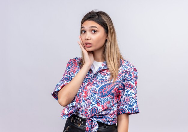 A surprising beautiful young woman wearing paisley printed shirt keeping hand on face