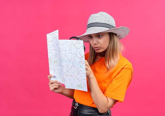 Free photo a surprising pretty young woman in an orange t-shirt wearing sunhat looking at a map with magnifying glass