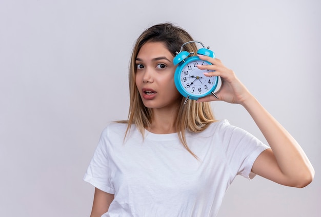A surprising young woman in white t-shirt listening to clock ticking sound while holding blue alarm clock