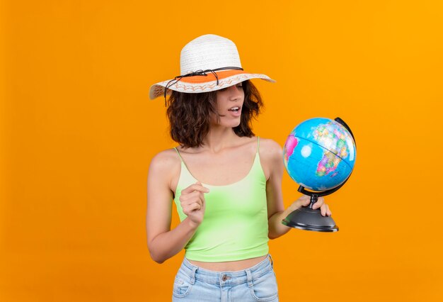 A surprising young woman with short hair in green crop top wearing sun hat looking at a globe 