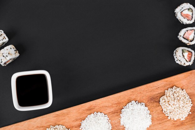 Sushi rolls; soya sauce and uncooked rice on wooden tray over the black background