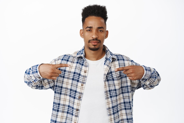 Free Photo suspicious african american man squinting doubtful, pointing fingers at center, showing logo or banner at center, frowning upset, standing against white background