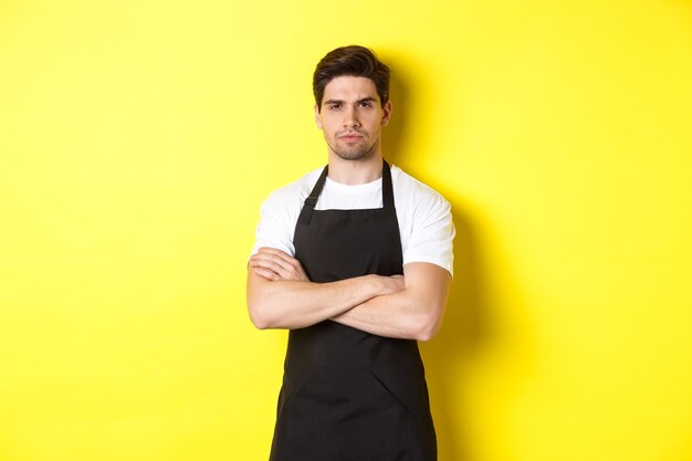 Suspicious barista squinting, cross arms on chest and looking at something with disbelief, standing over yellow background.