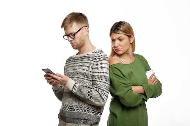 Free photo suspicious distrustful young blonde female in green top standing next to her bearded boyfriend, looking over his shoulder, spying over him while he is typing message using mobile phone