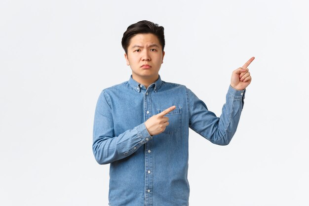 Suspicious and doubtful asian male in blue shirt, frowning skeptical and displeased, pointing fingers upper right corner, asking question, complaining on something bothering, white background.