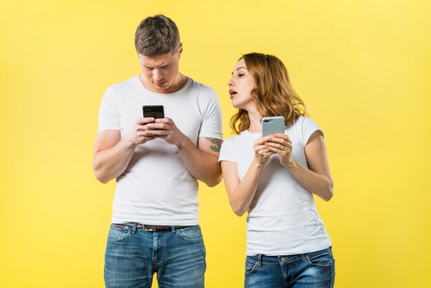 Suspicious girlfriend spying his boyfriend texting on mobile phone against yellow backdrop