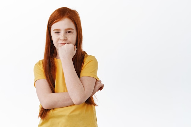Suspicious little redhead girl having doubts, looking with disbelief and hesitant face at camera, thinking, standing over white background