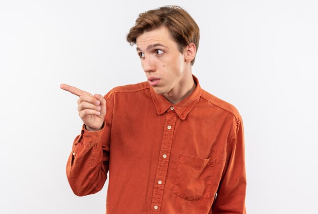 Suspicious looking side young handsome guy wearing red shirt points at side isolated on white wall