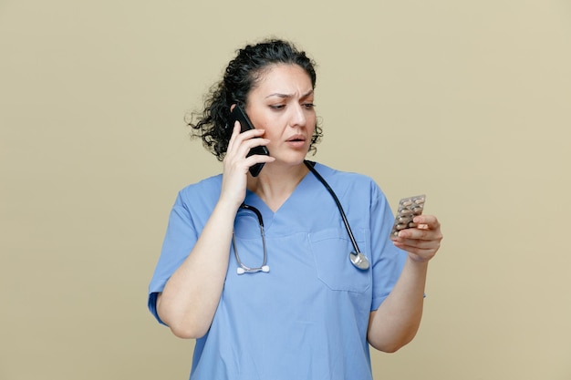 의심스러운 중년 여성 의사는 제복을 입고 목에 청진기를 두르고 올리브 배경에 격리된 팩을 보며 전화 통화를 하는 알약 팩을 들고 있습니다.