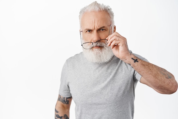 Suspicious old man with tattoos, take off glasses and looking with disbelief and angry face, confused about something strange, standing against white wall
