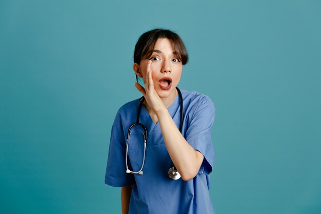 подозрительно показывая уверенную молодую женщину-врача в униформе, стетоскоп, изолированный на синем фоне
