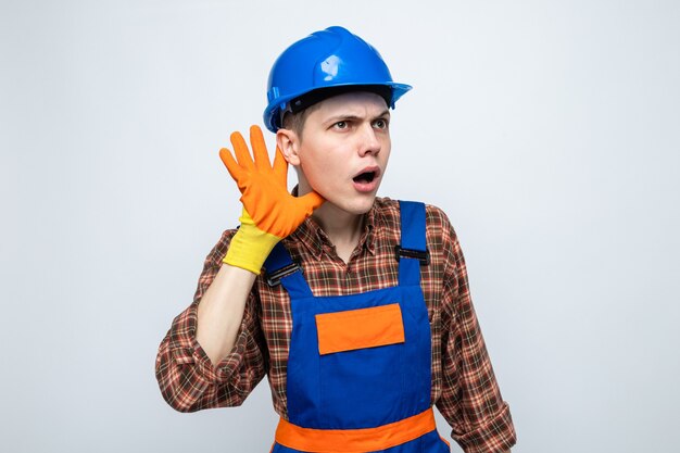 Suspicious showing listen gesture young male builder wearing uniform with gloves 