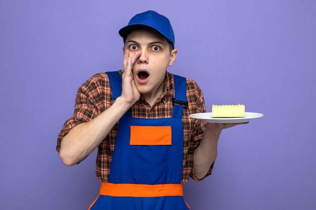 Suspicious whispers young cleaning guy wearing uniform and cap holding sponge on plate 