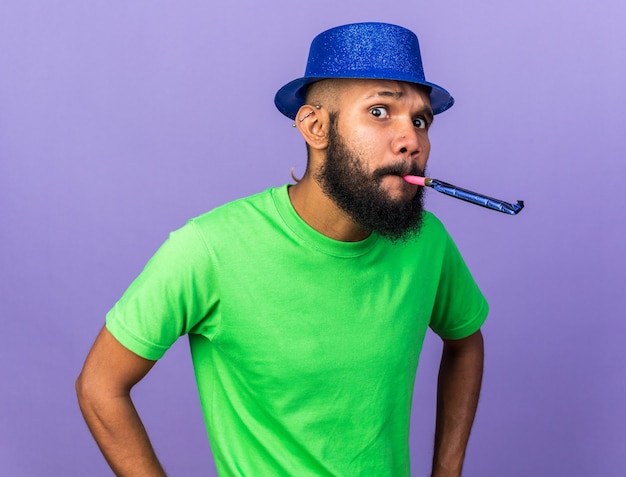 Suspicious young afro-american guy wearing party hat blowing party whistle