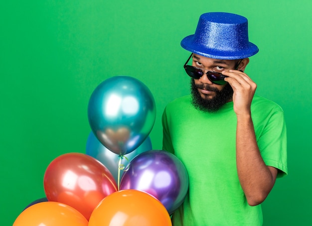 Free photo suspicious young afro-american guy wearing party hat and glasses holding balloons