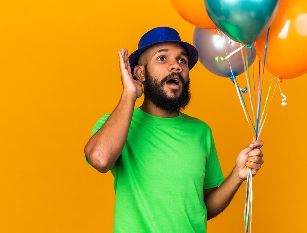 Suspicious young afro-american guy wearing party hat holding balloons showing listen gesture 